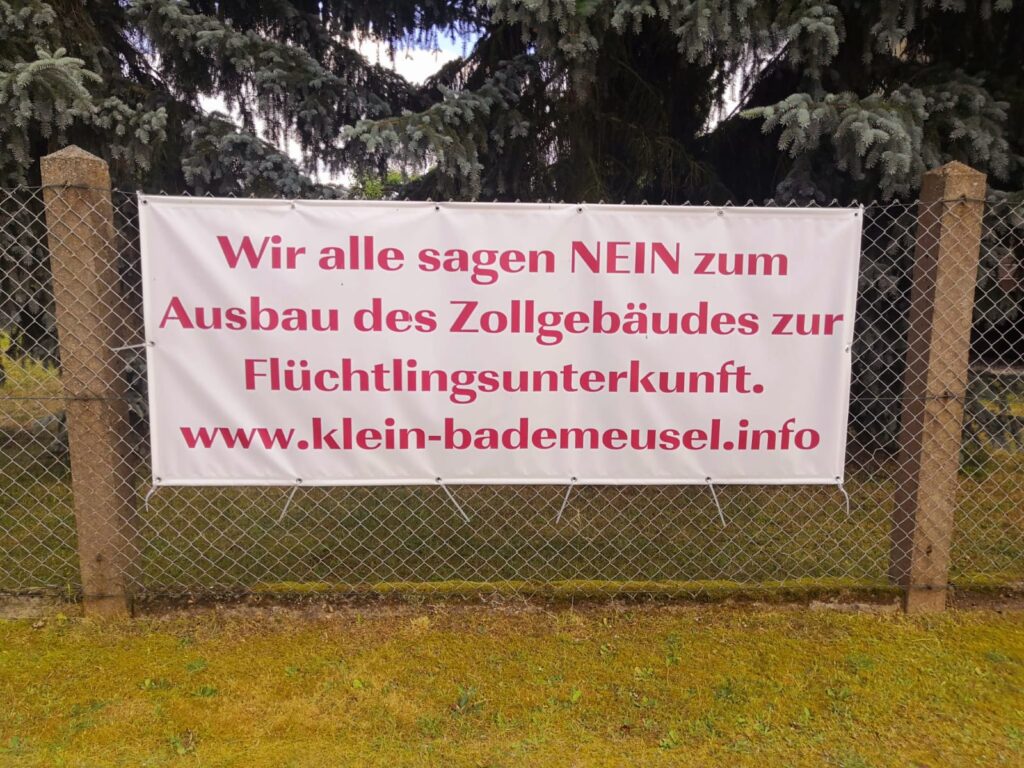 Poster an einem Zaun, mit dem Text: "Wir alle sagen Nein zum Ausbau des Zollgebäudes zur Flüchtlingsunterkunft. www.klein-bademeusel.info"