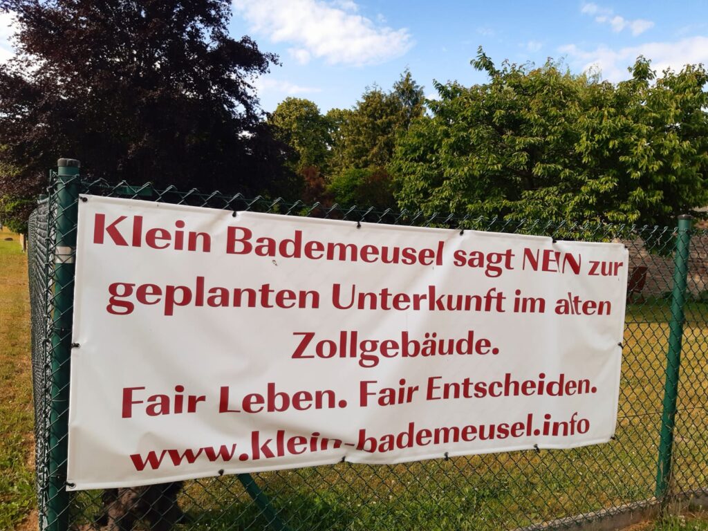 Großes Plakat an einem Gartenzaun, mit dem Text: "Klein Bademeusel sagt Nein zur geplanten Unterkunft im alten Zollgebäude. Fair Leben. Fair Entscheiden. www.klein-bademeusel.info"