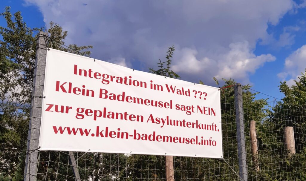 Großes Plakat an einem Zaun, mit dem Text: "Integration im Wald? Klein Bademeusel sagt Nein zur geplanten Asylunterkunft. www.klein-bademeusel.info"